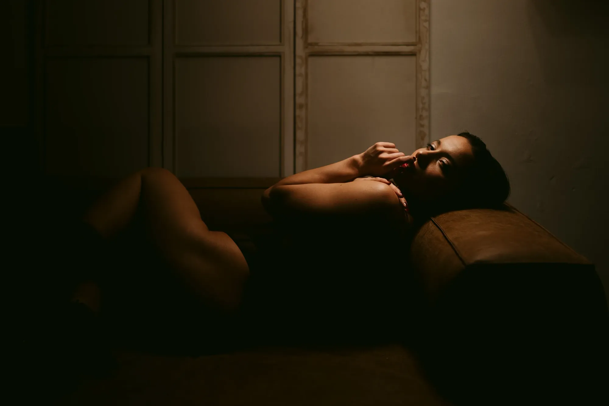 A moody, low-light portrait of woman reclining on a brown sofa, wearing dark lingerie, with warm light highlighting her face and legs, taken by Boudoir Photographer Chris Zielecki