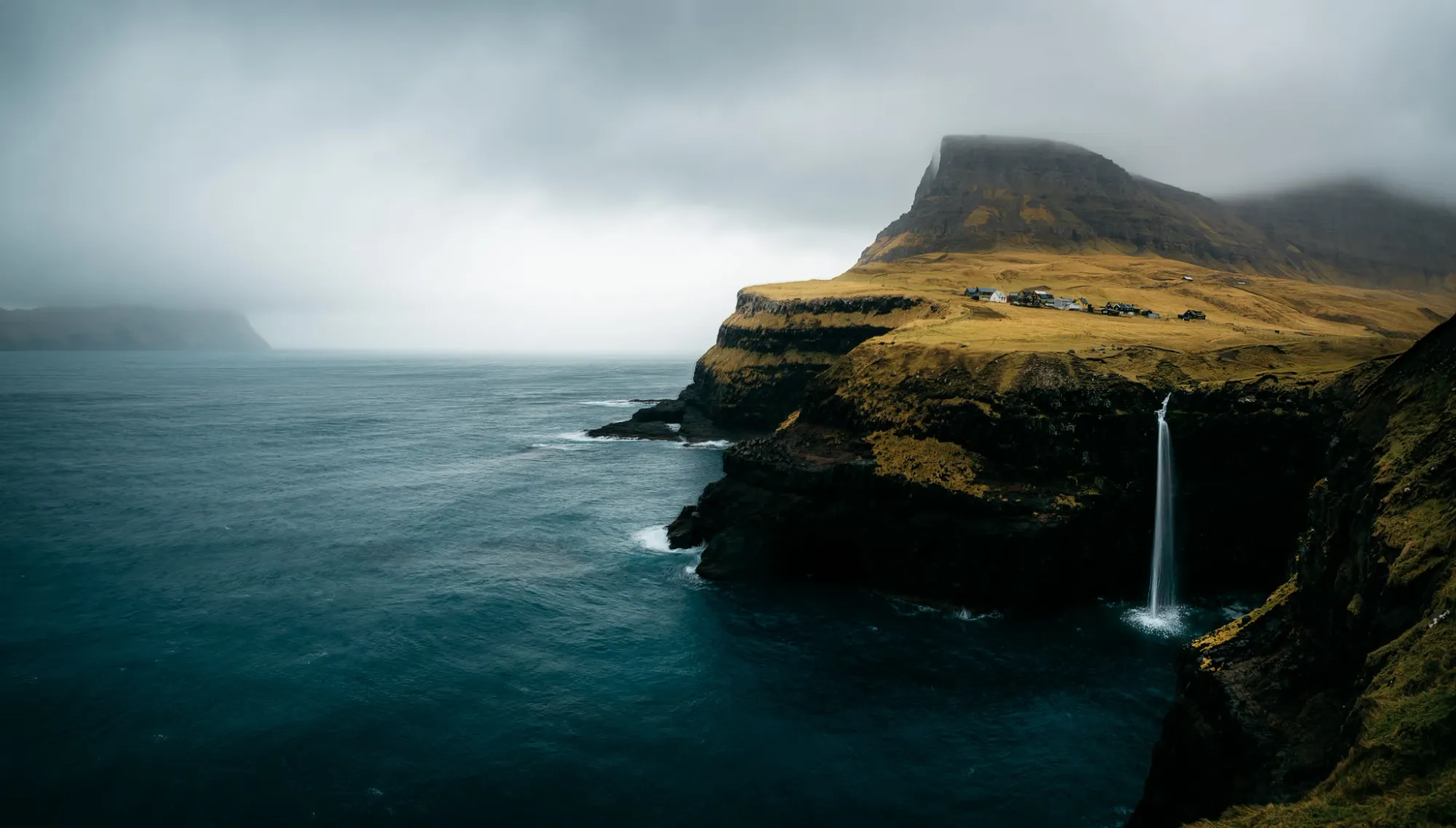 A remote coastal cliff with a tall waterfall plunging into a dark blue ocean, topped by a small village under a cloudy, misty sky, captured by Chris Zielecki at the Faroe Islands Photo Tour